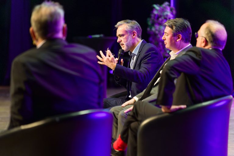 Quatre hommes en costume sont assis sur des chaises noires et participent à une table ronde sur le DOMOFRANCE. L'un d'eux, au centre, parle dans un microphone en faisant un geste de la main. L'arrière-plan est sombre, avec un éclairage de scène violet et une plante feuillue visible derrière.