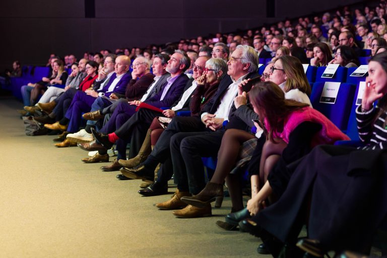 Dans un auditorium faiblement éclairé, un large public d'hommes et de femmes, souvent en tenue de ville ou d'affaires, est assis sur des rangées de chaises bleues et regarde attentivement la présentation de DOMOFRANCE sur scène, avec des expressions concentrées et engagées.