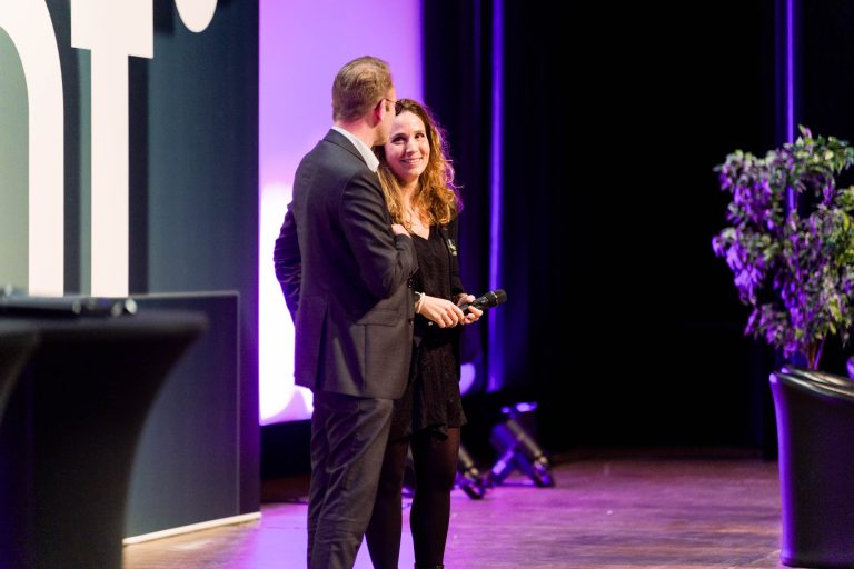 Une femme en robe noire tenant un micro se tient sur scène, souriant à un homme en costume. Ils sont sous un éclairage violet, près de grandes lettres blanches DOMOFRANCE sur le mur, avec des plantes en pot et des éléments d'arrière-plan flous.