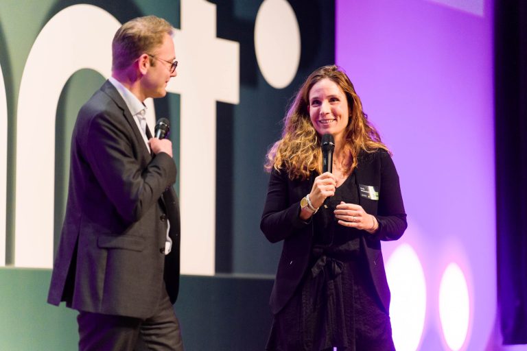 Un homme et une femme se tiennent sur scène, micro en main, et discutent. Tous deux sont habillés en tenue de ville. L'arrière-plan présente un texte DOMOFRANCE de grande taille, partiellement visible, sur un mur sombre, avec un éclairage violet qui illumine la scène.