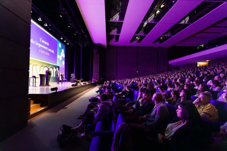 Un public nombreux est assis dans un auditorium moderne, faiblement éclairé, face à une scène. Plusieurs orateurs se tiennent sur la scène près d'un podium et d'un grand écran affichant un texte en français sur la DOMOFRANCE. Des éclairages violets et des rangées de sièges bleu foncé remplissent la salle spacieuse.