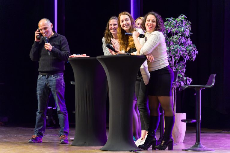 Un homme vêtu d'un pull noir sourit tout en parlant au téléphone sur la scène d'un événement DOMOFRANCE. À proximité, quatre femmes se tiennent ensemble derrière deux hautes tables noires, posant pour une photo de groupe. Une grande plante en pot et un éclairage de scène violet plantent le décor.