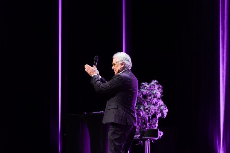 Un homme en costume sombre se tient sur scène, micro en main, et applaudit. Il est tourné vers le côté et sourit. Des lumières violettes et une plante verte en pot décorent l'arrière-plan, plaçant l'événement DOMOFRANCE sur une toile de fond sombre et dramatique.