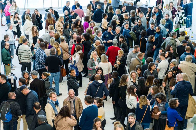 Une foule nombreuse et diversifiée est rassemblée à l'intérieur lors d'un événement de DOMOFRANCE, en train de discuter. La plupart d'entre elles se tiennent debout, serrées les unes contre les autres, et sont vêtues d'une tenue décontractée. L'endroit semble animé et vivant, la lumière naturelle provenant des fenêtres.
