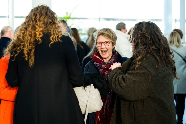 Une femme aux cheveux courts et aux lunettes sourit tout en discutant avec deux autres personnes portant des manteaux d'hiver lors d'un événement intérieur très fréquenté de DOMOFRANCE. Elle porte un foulard et un manteau sombre. L'arrière-plan est rempli de personnes en conversation et la lumière naturelle y pénètre.