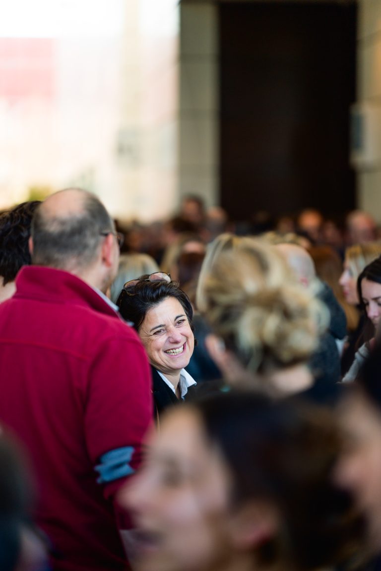 Une femme aux cheveux courts et bruns sourit avec éclat dans un espace intérieur bondé lors d'un événement DOMOFRANCE. Elle se tient au milieu de nombreuses personnes, dont certaines sont tournées vers l'extérieur. La scène animée est éclairée par une lumière naturelle et des silhouettes légèrement floues à l'arrière-plan.