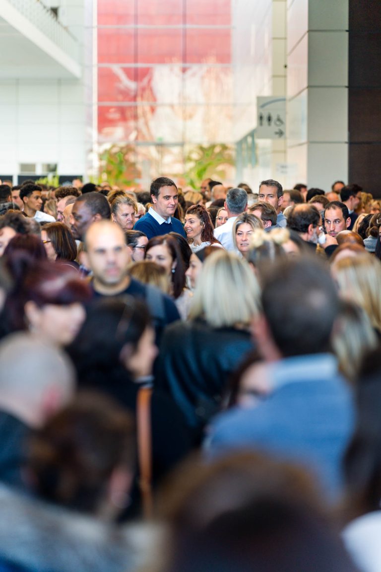Une grande foule de personnes se tient serrée les unes contre les autres à l'intérieur, remplissant un couloir spacieux d'une lumière vive chez DOMOFRANCE. La plupart des personnes sont tournées vers l'avant ou sur le côté, beaucoup sont engagées dans une conversation, ce qui crée une atmosphère animée.