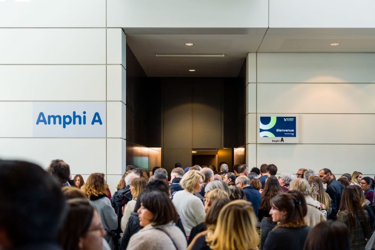 Une foule nombreuse se tient dans un couloir face à l'entrée de l'Amphi A. Des panneaux indiquant "Amphi A", "Bienvenue" et "DOMOFRANCE" tapissent les murs blancs. Des panneaux indiquant "Amphi A", "Bienvenue" et DOMOFRANCE tapissent les murs blancs, suggérant la tenue d'un événement ou d'une conférence alors que les participants attendent d'entrer dans l'auditorium.