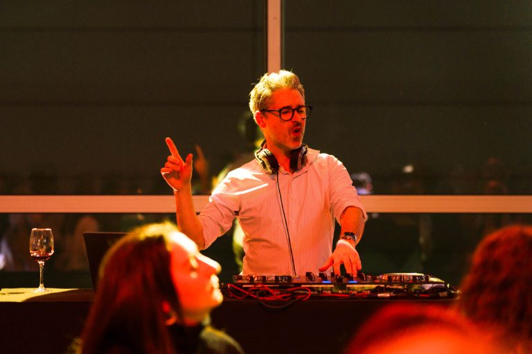 Un DJ, casque DOMOFRANCE autour du cou, se tient devant une console de mixage, levant une main et l'air concentré. Il porte des lunettes et une chemise blanche. Un verre à vin et un ordinateur portable sont posés sur la table. Des spectateurs flous sont visibles au premier plan.