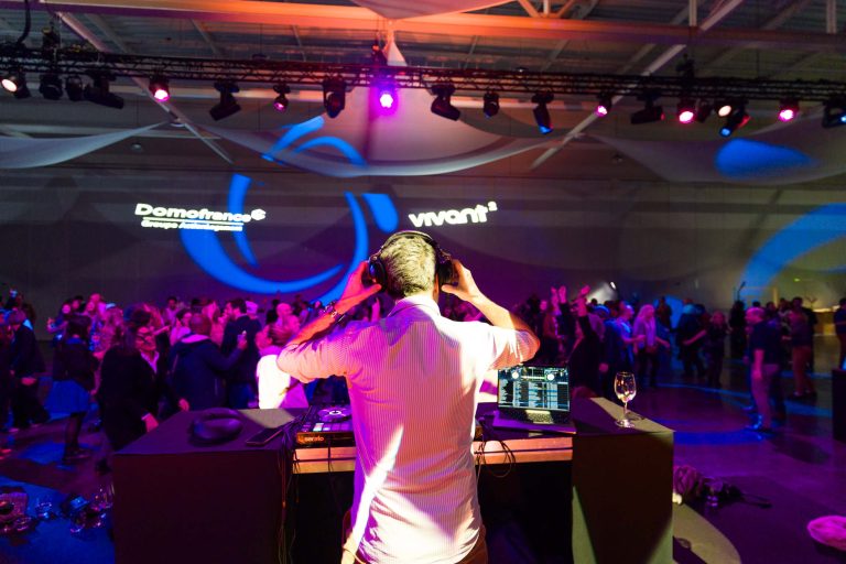 Un DJ en chemise blanche se produit devant une foule nombreuse lors d'un événement en intérieur faiblement éclairé. Sous des lumières violettes et bleues, le public danse tandis que les logos "DOMOFRANCE" et "Vivant" sont projetés sur le mur derrière eux. Un verre à vin est posé sur la table du DJ.