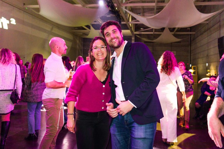 Un homme souriant vêtu d'un blazer sombre et une femme portant des lunettes et un pull-over magenta posent ensemble pour une photo lors d'une fête DOMOFRANCE animée à l'intérieur, avec des gens qui se mélangent, des lumières colorées et des rideaux de tissu blanc au plafond.