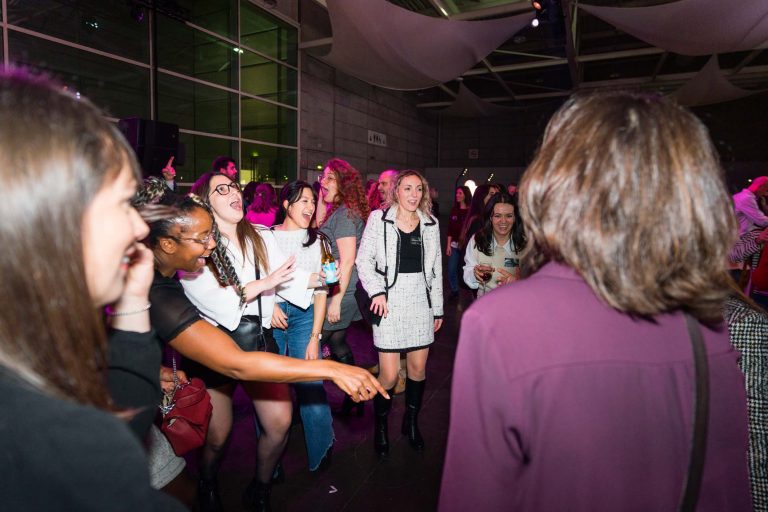Un groupe de jeunes femmes, dont certaines rient et pointent du doigt, dansent ensemble lors d'un événement DOMOFRANCE en intérieur, sous un éclairage rose et violet. Elles portent des tenues semi-formelles. Les hauts plafonds et les tissus drapés ajoutent à l'atmosphère festive.