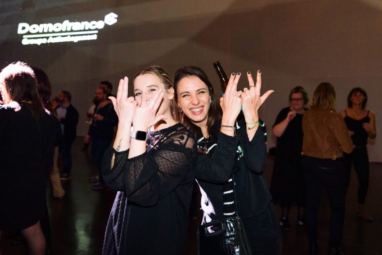 Deux femmes en tenue noire sourient et posent en faisant des gestes ludiques de la main lors d'un événement animé en intérieur. D'autres invités se mêlent à la foule tandis que le logo lumineux de DOMOFRANCE est projeté sur le mur, soulignant l'atmosphère sociale.