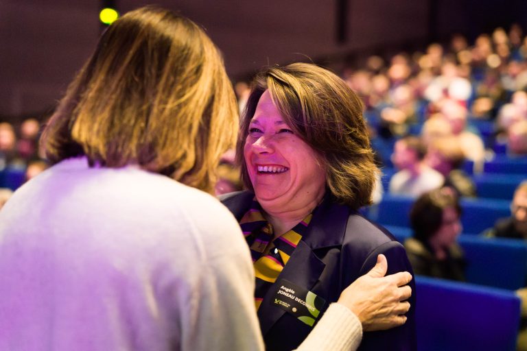 Deux femmes échangent chaleureusement dans un auditorium bondé. L'une, face à la caméra, sourit largement dans son badge DOMOFRANCE et son blazer sombre ; l'autre, vue de dos, pose une main sur son épaule. Des spectateurs flous occupent les sièges bleus à l'arrière-plan.