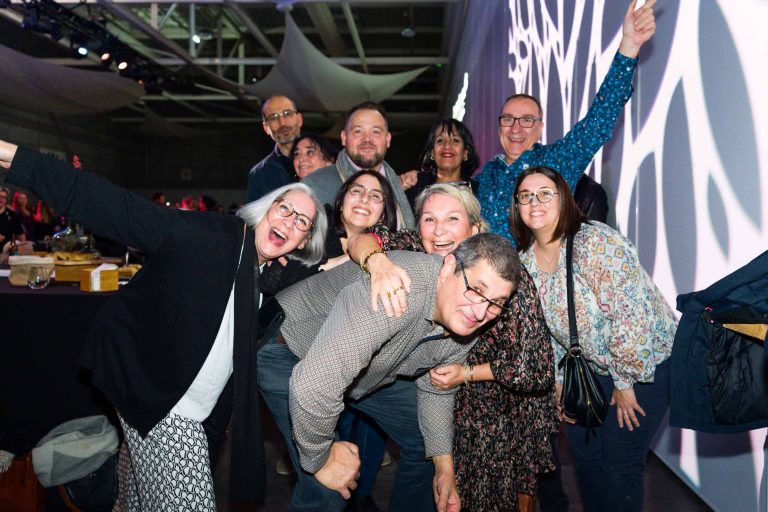 Un groupe de dix adultes pose étroitement ensemble lors d'un événement DOMOFRANCE en salle, en souriant et en riant. Certaines personnes ont les bras levés ou s'entourent. L'arrière-plan montre un éclairage dramatique et des motifs lumineux géométriques sur le mur.