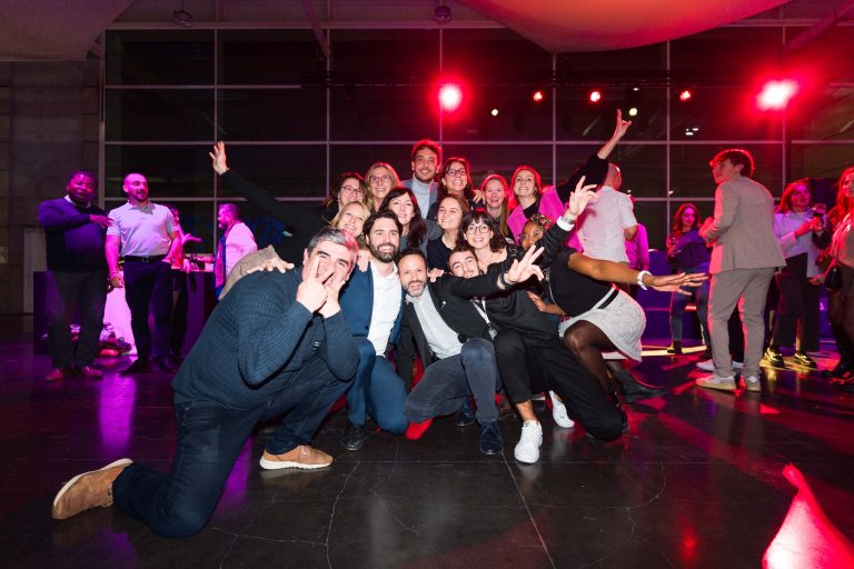 Un groupe de personnes sourit et pose énergiquement ensemble lors d'une fête DOMOFRANCE, en s'accroupissant et en faisant des gestes ludiques. À l'arrière-plan, d'autres personnes discutent sous un éclairage violet et rouge dans une salle intérieure moderne dotée de grandes fenêtres. L'ambiance est joyeuse et festive.