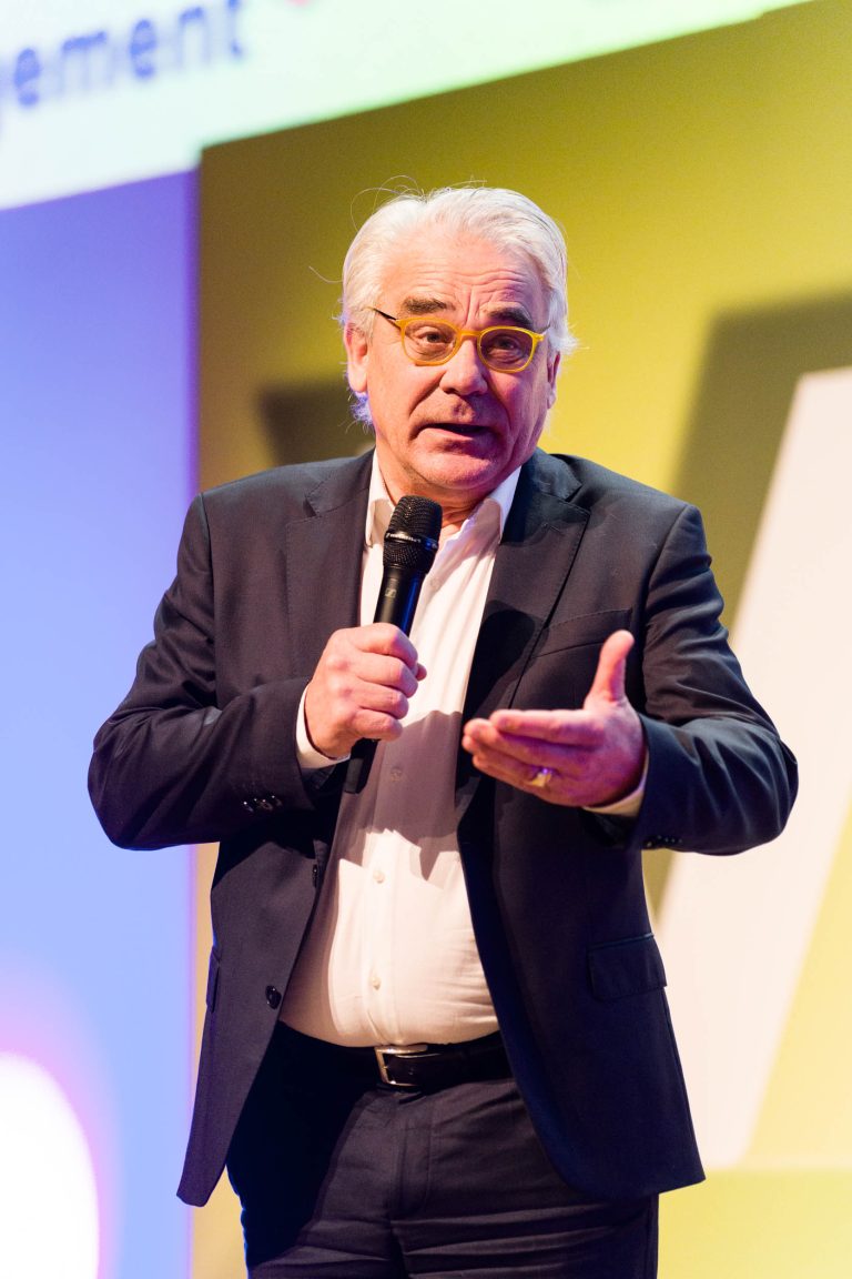 Un homme âgé aux cheveux blancs, portant des lunettes, un costume sombre et une chemise blanche, tient un micro d'une main et fait des gestes tout en parlant sur une scène DOMOFRANCE brillamment éclairée sur fond jaune et violet.