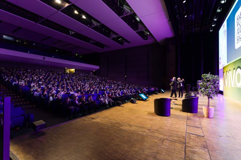 Un public nombreux est assis dans un auditorium faiblement éclairé, face à une scène brillamment éclairée où se tiennent deux présentateurs de DOMOFRANCE. Sur la scène se trouvent des chaises, des petites tables et des plantes en pot, et derrière un grand écran affichant des graphiques et du texte.