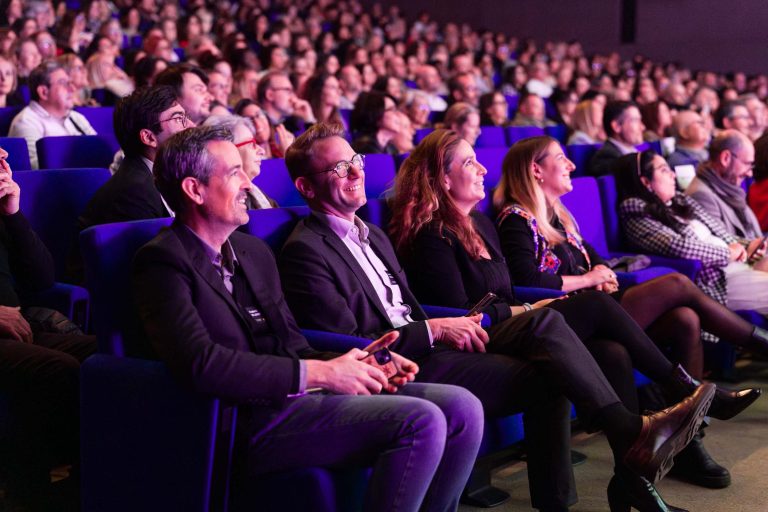 Un public nombreux est assis sur des rangées de chaises bleues lors d'un événement DOMOFRANCE en salle. Des personnes en tenue de travail sourient et regardent la scène avec intérêt. La salle remplie et chaleureusement éclairée crée une atmosphère vivante et attentive.