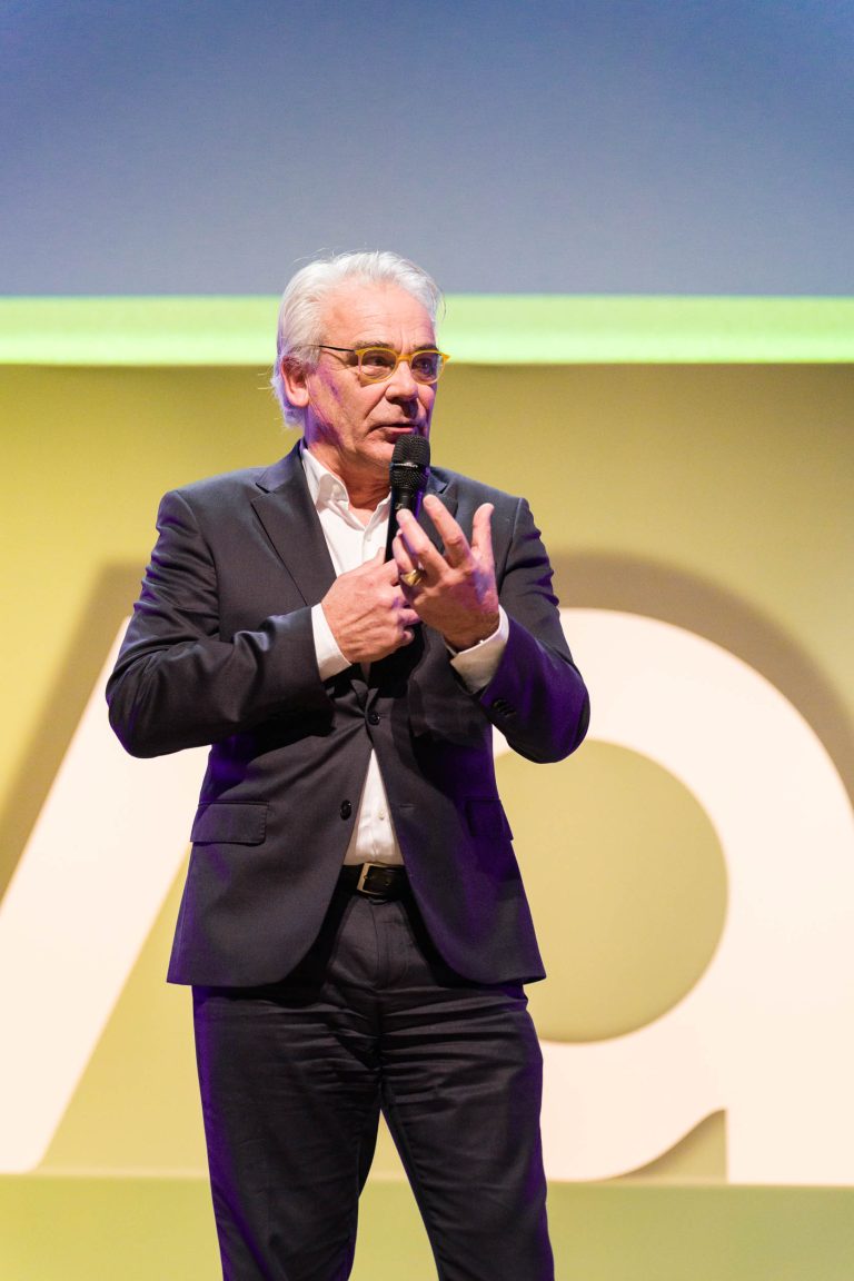 Un homme d'un certain âge, aux cheveux blancs, portant des lunettes et vêtu d'un costume sombre, tient un micro alors qu'il s'exprime sur scène lors d'un événement organisé par DOMOFRANCE. Il fait des gestes d'une main, une grande lettre blanche "A" et un fond jaune-vert étant visibles derrière lui dans ce cadre formel.