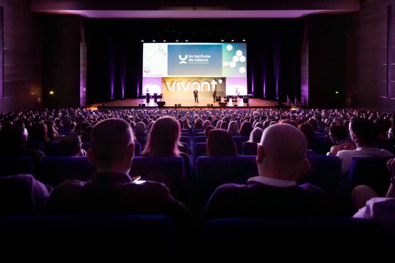 Un public nombreux est assis dans un auditorium sombre, face à une scène très éclairée où se trouvent plusieurs orateurs. L'écran derrière la scène affiche "VIVANT", "Un territoire de valeurs" et le logo de DOMOFRANCE au milieu de graphiques abstraits, créant ainsi une atmosphère de conférence formelle.