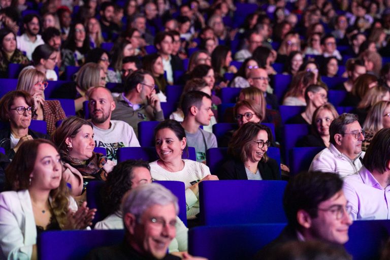 Un large public est assis sur des rangées de chaises bleues lors d'un événement DOMOFRANCE en salle. Des personnes d'âges divers, principalement des adultes, discutent, sourient et applaudissent. L'atmosphère animée est vibrante et sociale car tout le monde est tourné vers l'avant.