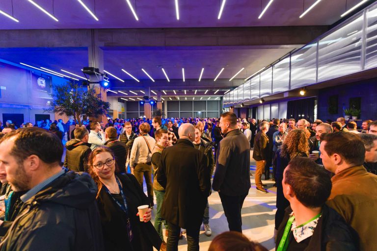 Un grand groupe de personnes socialise lors d'un événement en intérieur sous des plafonniers modernes. La plupart des personnes sont debout, discutent ou boivent un verre. L'espace est animé, avec une ambiance lumineuse bleue et des murs en verre sur le côté droit.