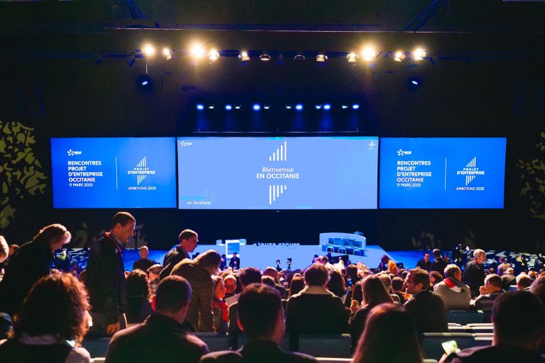 Un public nombreux se rassemble dans un auditorium faiblement éclairé, face à une large scène sur laquelle se trouvent trois écrans bleus sur lesquels on peut lire "Bienvenue en Occitanie". Les gens se lèvent, parlent et se préparent à s'asseoir. La scène est préparée pour un événement d'entreprise ou d'affaires, avec quelques tables et chaises.