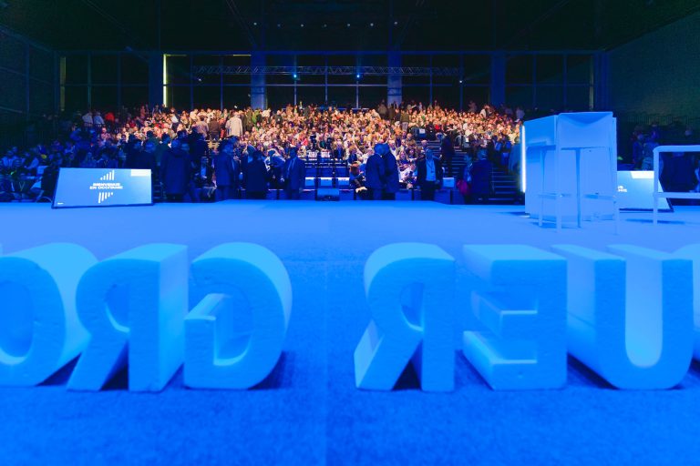 Un large public est assis sur des sièges en gradins lors d'un événement en intérieur, éclairé par une lumière bleue. Au premier plan, de grandes lettres blanches épellent un texte inversé sur le sol. Deux écrans affichent des graphiques et un mobilier moderne minimal se trouve sur la scène.