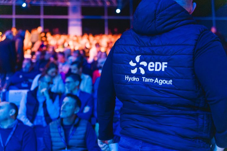 Une personne portant une veste sombre avec le logo "EDF Hydro Tarn-Agout" se tient face à un public assis dans un espace intérieur très éclairé et bondé. L'arrière-plan est rempli de personnes et éclairé par des lumières bleues et orange.