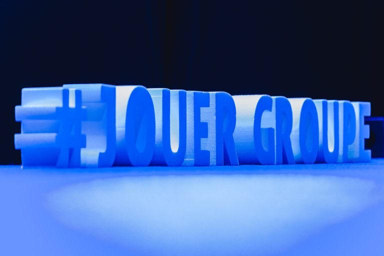 Une sculpture bleue en 3D du hashtag indique "#JOUER GROUPE" sur une surface plane et sur un fond sombre. Les lettres majuscules et le symbole apparaissent illuminés d'une teinte bleue, donnant à la scène un aspect moderne et saisissant.