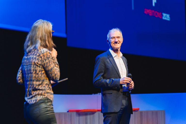 Un homme en costume sombre et chemise blanche sourit en tenant un micro sur scène. Une femme aux cheveux longs, aux lunettes et au haut à motifs lui fait face et parle. L'arrière-plan est bleu avec une projection abstraite et des meubles modernes.