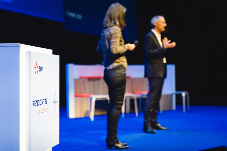 Sur une estrade bleue, deux personnes parlent ; une femme en pantalon de cuir tient un micro tandis qu'un homme en costume fait des gestes. Au premier plan, on aperçoit un podium avec le logo EDF et "RENCONTRE en Occitanie". L'arrière-plan montre des bancs et une toile de fond sombre.
