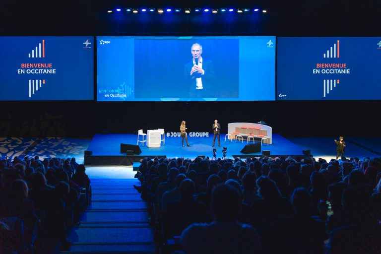Un public nombreux fait face à une scène où deux présentateurs s'expriment sous des lumières vives. Derrière eux, un écran affiche un homme et indique "Bienvenue en Occitanie". La scène est équipée de chaises, d'un podium et d'un décor bleu, créant ainsi une atmosphère de conférence formelle.