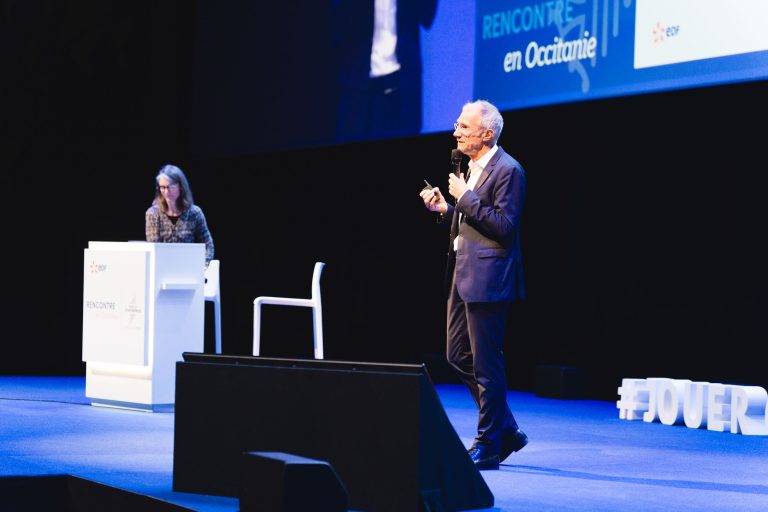 Un homme en costume parle sur scène en tenant un micro et une télécommande, tandis qu'une femme est assise sur un podium blanc à l'arrière-plan. La scène a un sol bleu, un grand écran et le hashtag "#JOVER" en lettres blanches sur la droite.