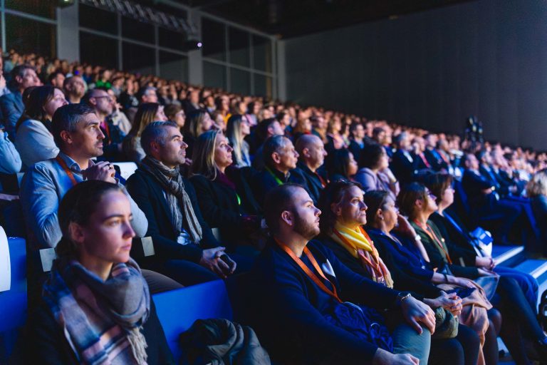 Un large public d'hommes et de femmes est assis en rangs serrés dans un auditorium faiblement éclairé, face à une scène brillamment éclairée. De nombreuses personnes portent des vêtements de travail et des cordons, ce qui indique qu'il s'agit d'une conférence ou d'un événement formel.