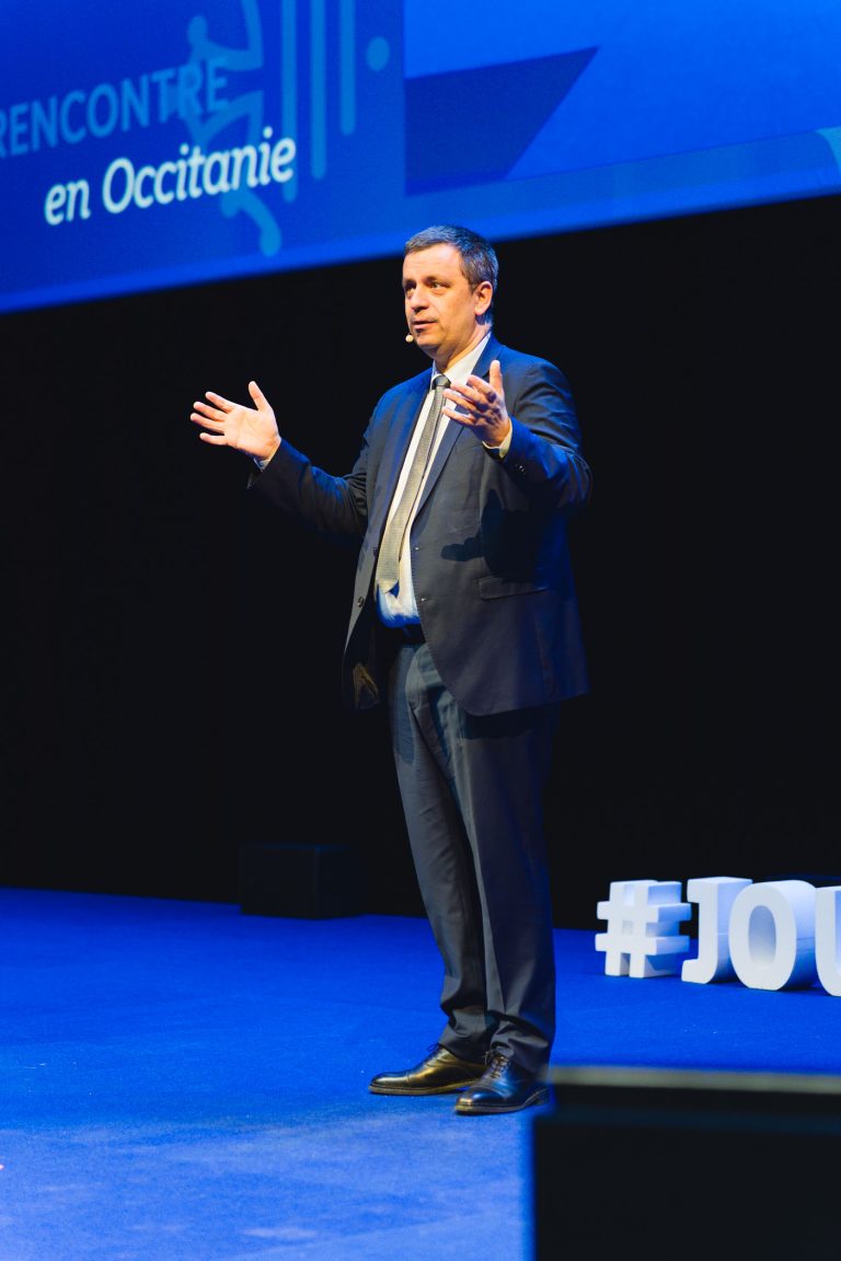 Un homme en costume sombre et cravate se tient sur une scène éclairée en bleu, les bras ouverts, en plein discours. Derrière lui, un grand écran affiche des textes et des graphiques en français. Des lettres blanches en 3D sur la scène épellent en partie "#JOU". Le cadre est formel, il s'agit probablement d'une conférence ou d'une présentation.