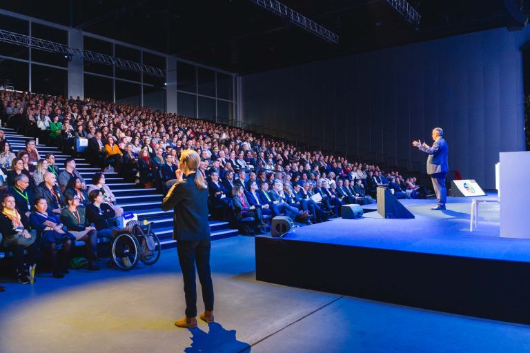 Un homme s'adresse sur scène à un large public assis dans un auditorium. Un interprète en langue des signes se tient à l'avant. Un membre du public se déplace en fauteuil roulant. La salle est très éclairée, les sièges sont disposés en gradins et les plafonds sont hauts.