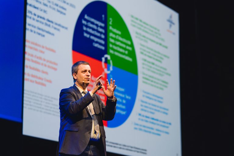Un homme en costume, équipé d'un micro-casque, fait des gestes pendant qu'il parle sur scène. Derrière lui, un grand écran affiche un tableau coloré et segmenté avec du texte, illustrant probablement les points clés de sa présentation. L'arrière-plan est sombre, concentrant l'attention sur l'orateur et l'écran.
