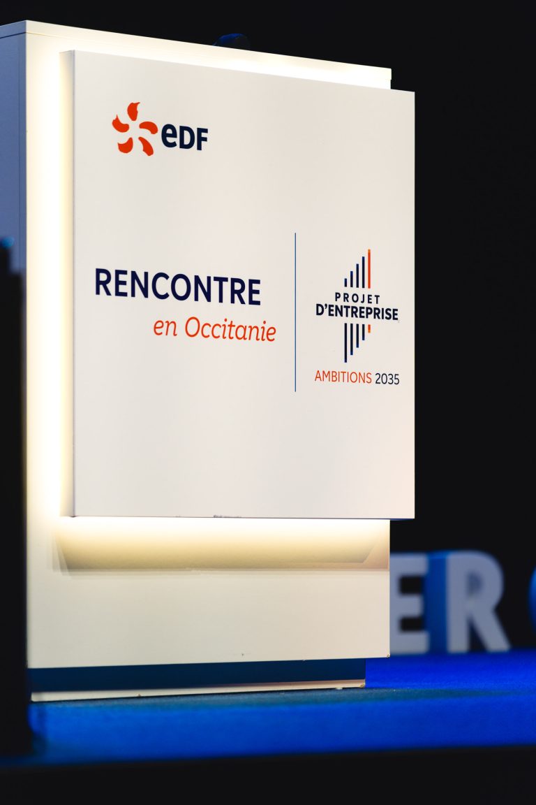 Un podium blanc affiche le logo EDF et le texte "RENCONTRE en Occitanie", ainsi que "PROJET D'ENTREPRISE AMBITIONS 2035" sur la face avant, avec des accents bleus et orange, sur un fond sombre.