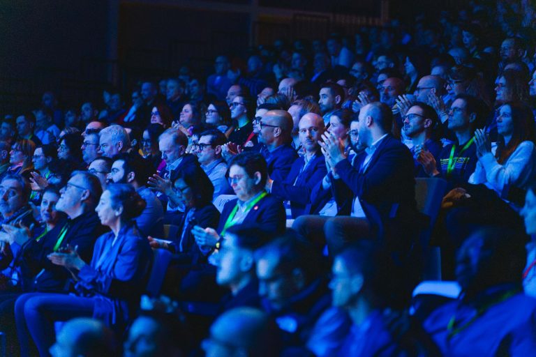Un public nombreux est assis, serré les uns contre les autres, dans un auditorium faiblement éclairé par une lumière bleue. De nombreuses personnes applaudissent, d'autres regardent la scène et quelques-unes utilisent leur téléphone. L'ambiance semble engagée et attentive.