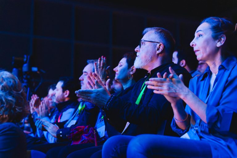 Un groupe de personnes est assis dans un public faiblement éclairé, applaudissant et regardant vers la scène. Elles portent des vêtements d'affaires ou décontractés et semblent engagées et attentives, baignées dans un éclairage bleu et violet. L'arrière-plan est sombre et flou.