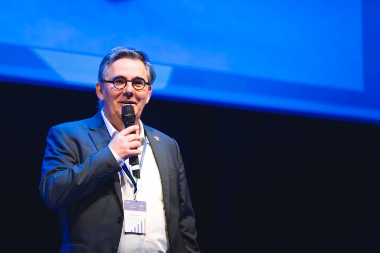 Un homme en costume et lunettes se tient sur scène et tient un microphone. Il porte un badge et sourit sous un éclairage de scène vif, avec un fond bleu derrière lui, suggérant qu'il fait une présentation ou un discours lors d'un événement ou d'une conférence.