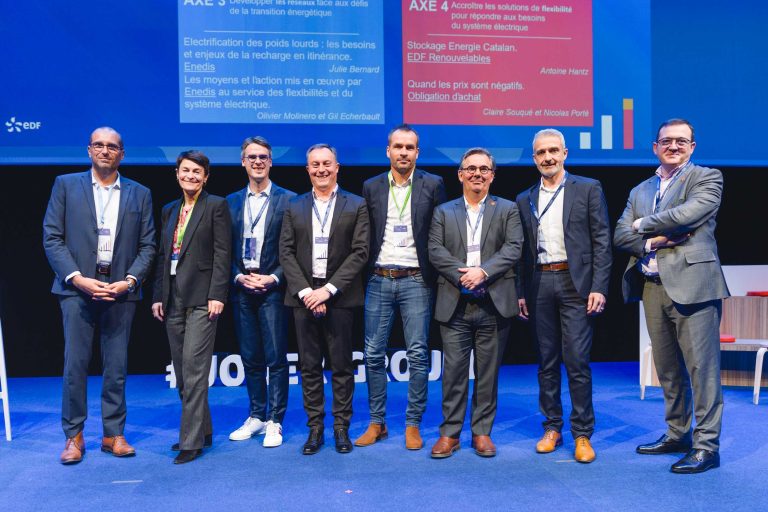 Huit personnes habillées professionnellement se tiennent en rang sur une scène bleue, souriant à la caméra. Derrière elles, un grand écran affiche des textes et des graphiques colorés relatifs à l'énergie et à la technologie. Deux chaises blanches sont visibles de chaque côté du groupe.