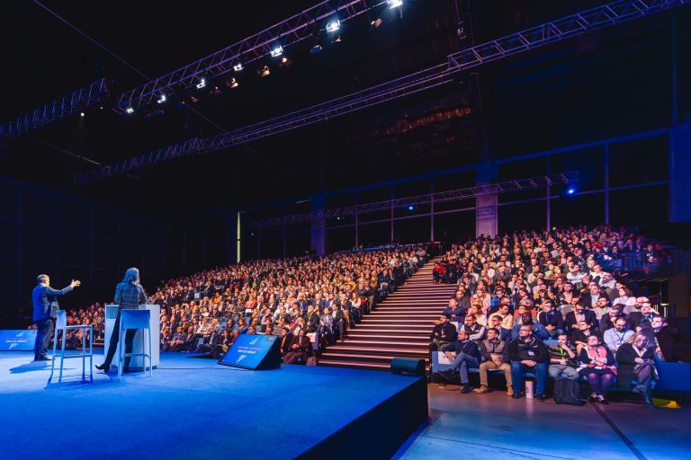 Deux orateurs sont assis sur la scène, face à un large public dans un auditorium moderne. La foule remplit les sièges en gradins sous un éclairage bleu et violet vif, et se concentre sur les présentateurs. Des écrans géants et des éclairages de scène encadrent le cadre professionnel de l'événement.