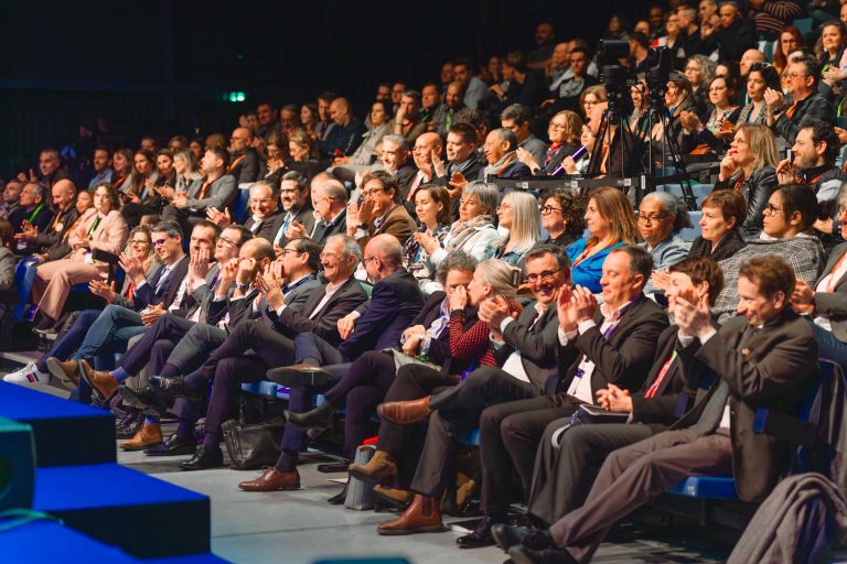 Un large public d'adultes divers, hommes et femmes, habillés en tenue de travail, est assis en rangs étagés lors d'un événement en salle. Certains applaudissent, d'autres parlent ou écoutent attentivement. L'éclairage est vif et l'atmosphère semble formelle et engagée.