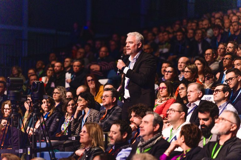 Un homme aux cheveux gris et vêtu d'un blazer noir se tient au milieu d'un large public assis et parle dans un microphone. La foule est variée, attentive et habillée en tenue de travail. Les cordons de l'événement et un appareil photo sur trépied sont visibles dans ce cadre vibrant et bien éclairé.