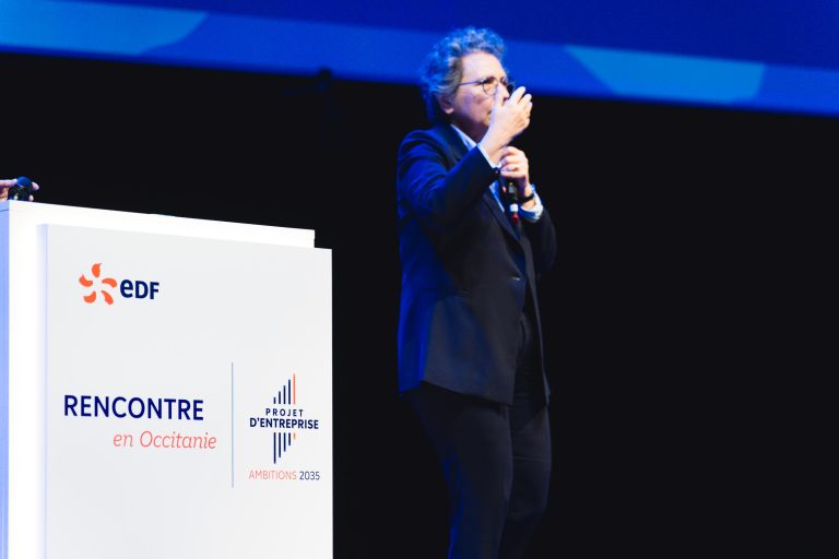Une personne en costume s'exprime sur scène à côté d'un podium blanc portant les inscriptions "EDF", "RENCONTRE en Occitanie" et "Projet D'Entreprise Ambitions 2035", faisant un geste d'une main tout en tenant un micro de l'autre, sur un fond sombre avec un éclairage bleu.