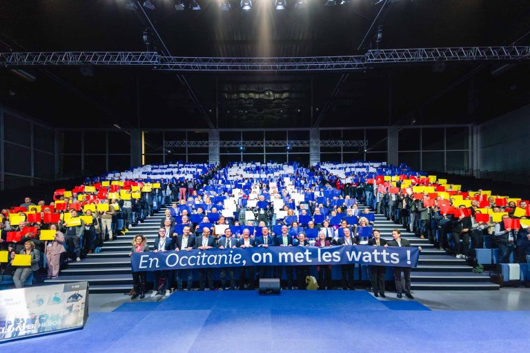 Un grand groupe de personnes est assis dans les gradins d'un forum, tenant des cartes rouges, jaunes et bleues pour former l'acronyme EDF. Devant, d'autres personnes tiennent une banderole sur laquelle on peut lire "En Occitanie, on met les watts !". Le décor est intérieur, avec des lumières de scène au-dessus et un premier plan recouvert d'une moquette bleue.