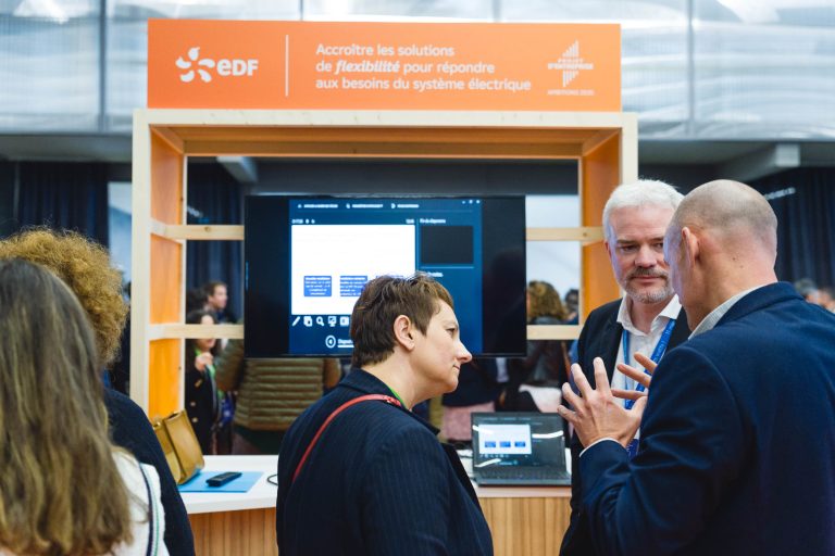 Trois personnes discutent devant une cabine orange portant le logo d'EDF et un texte en français sur la flexibilité du système électrique. Un écran affiche des diagrammes et du texte. D'autres participants sont visibles à l'arrière-plan de l'événement en salle.